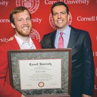 Two people stand in front of a red backgroun, holding a framed diploma