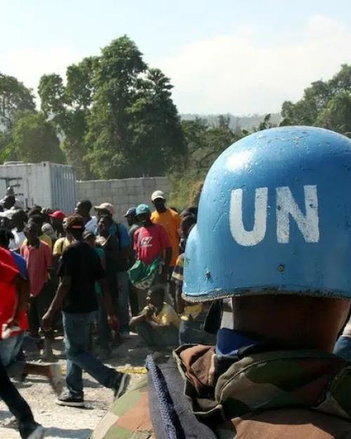 A UN blue peacekeeper's helmet in the foreground; facing a crowd of people