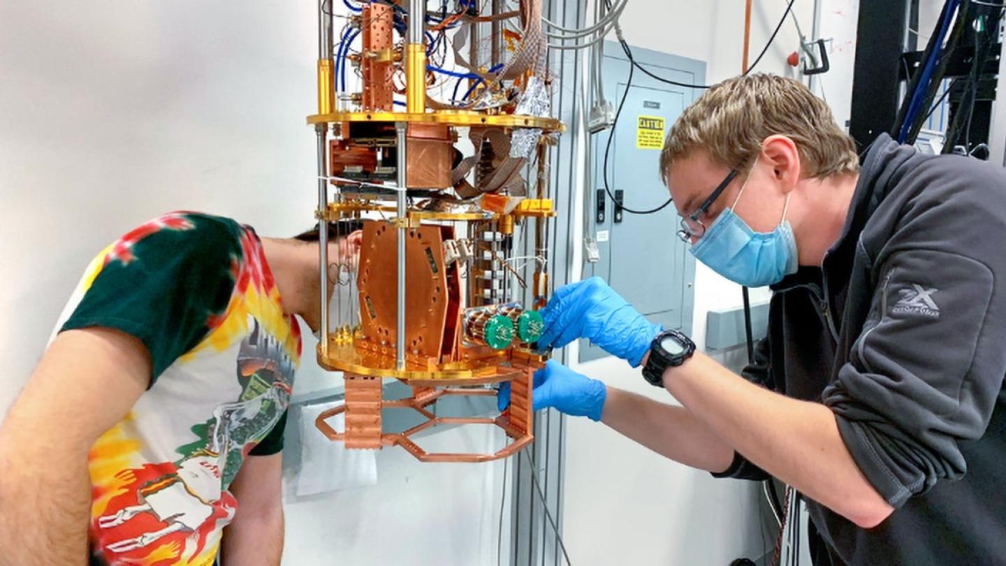 Two people lean toward a complicated scientific instrument featuring gold and orange metal parts; it's about the size of a coffeemaker